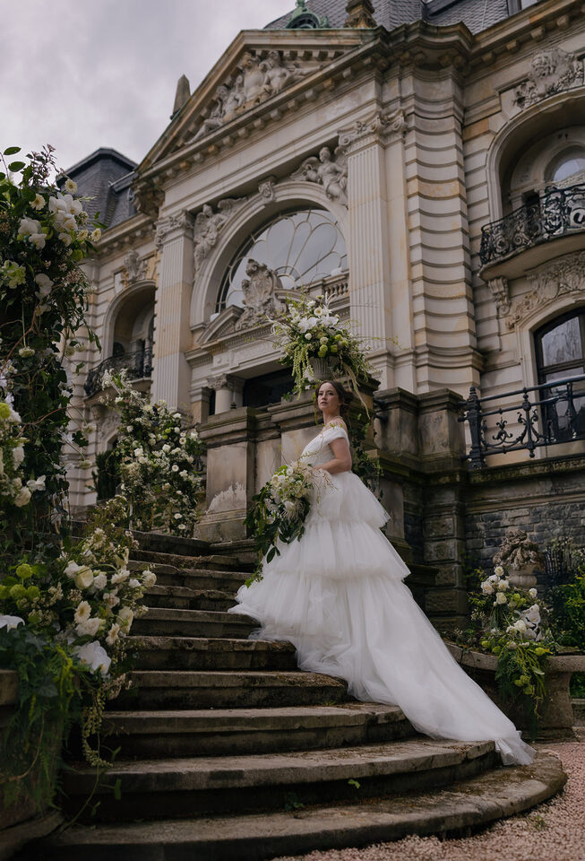 Eine Braut in einem voluminösen weißen Hochzeitskleid mit langer Schleppe steht auf der geschwungenen Steintreppe eines historischen Gebäudes. Sie hält einen großen locker gebundenen Brautstrauß aus weißen und grünen Blüten. Die Treppe ist auf beiden Seiten mit üppigen floralen Arrangements aus weißen Blumen, viel Grün und herabhängenden Zweigen dekoriert.