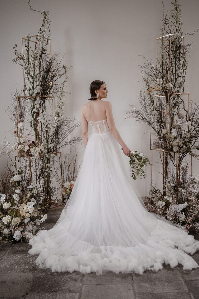 Eine Braut wird von hinten in einem langen weißen Tüllkleid mit großer Schleppe fotografiert. Sie steht zwischen zwei hohen, asymmetrischen floralen Installationen aus kahlen und geschwungenen Zweigen, weißen und cremefarbenen Blüten sowie einzelnen zarten Farbakzenten. Goldfarbene geometrische Metallrahmen sind in die Arrangements integriert. In einer Hand hält die Braut einen kleinen locker gebundenen Blumenstrauß.