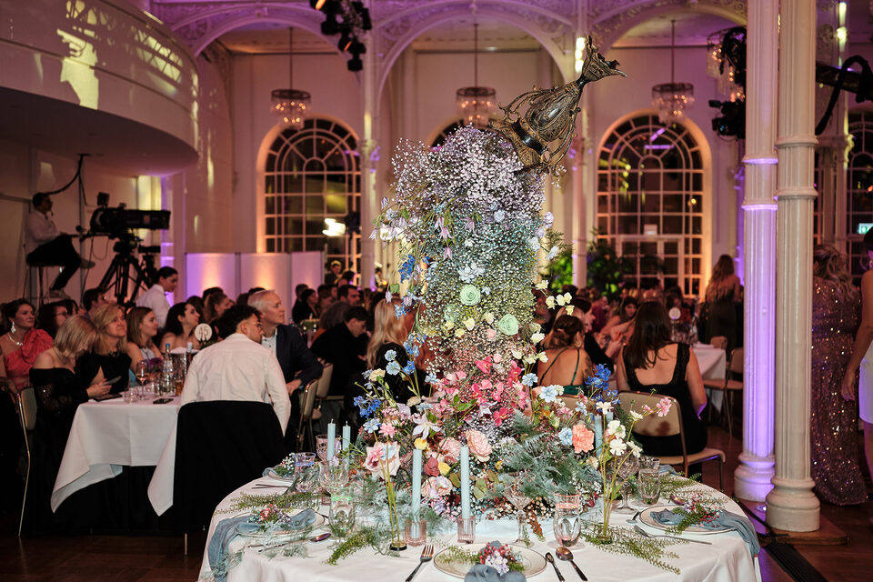 Eine festlich gedeckte runde Hochzeitstafel steht in einem großen historischen Saal mit hohen Rundbogenfenstern und violetter Beleuchtung. Auf die Tischmitte gießt sich eine mehrere Meter hohe, üppige Blumeninstallation mit Schleierkraut, rosa, weißen und blauen Blüten sowie viel Grün aus einer bronzenen Vase. Die Floristik reicht weit über die Köpfe der sitzenden Gäste hinaus.