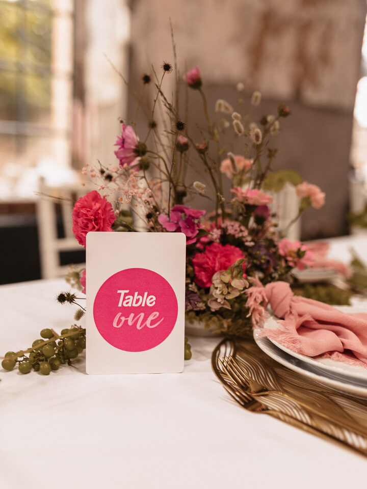 Auf einer Hochzeitstafel steht ein locker arrangiertes Blumengesteck mit kräftig pinken, rosafarbenen und einzelnen cremefarbenen Blüten, feinen Gräsern und langen Zweigen. Davor steht eine weiße Tischnummer mit einem großen pinken Kreis und der Aufschrift „Table one“. Rechts liegen rosa Stoffservietten auf gestapelten Tellern mit goldenem Besteck.