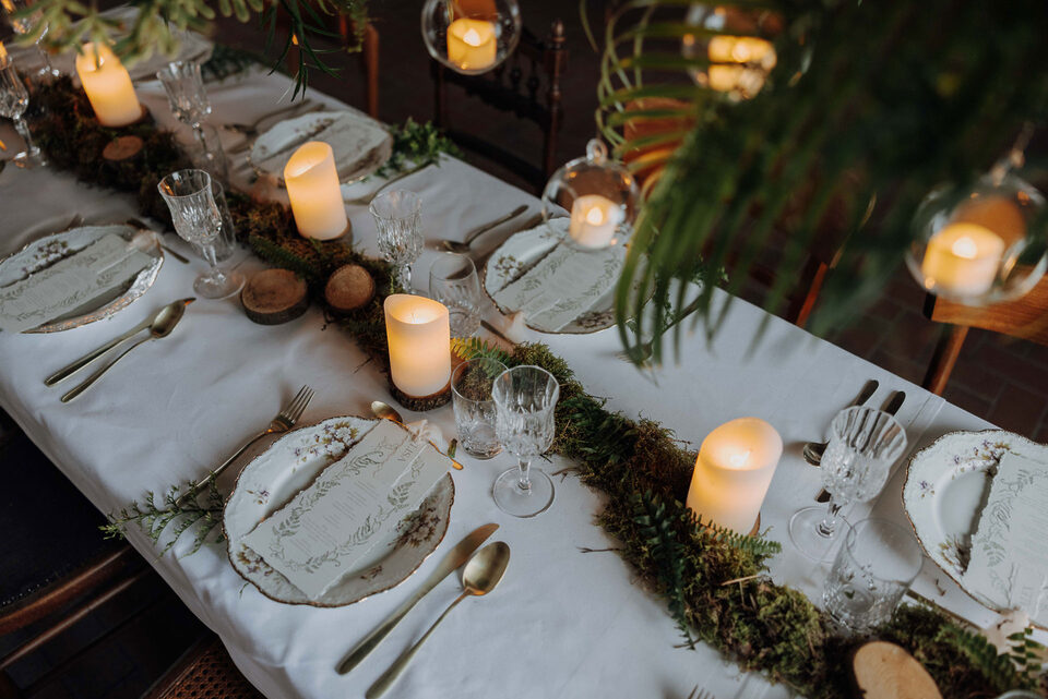 Eine festlich gedeckte Hochzeitstafel mit weißer Tischdecke, gemustertem Porzellan, Kristallgläsern und goldenem Besteck. Über die Tischmitte zieht sich eine üppige Dekoration aus Moos, Farnen, Grün und kleinen Holzscheiben. Dazwischen stehen unterschiedlich große leuchtende Kerzen; weitere Kerzen hängen in Glaskugeln über dem Tisch.