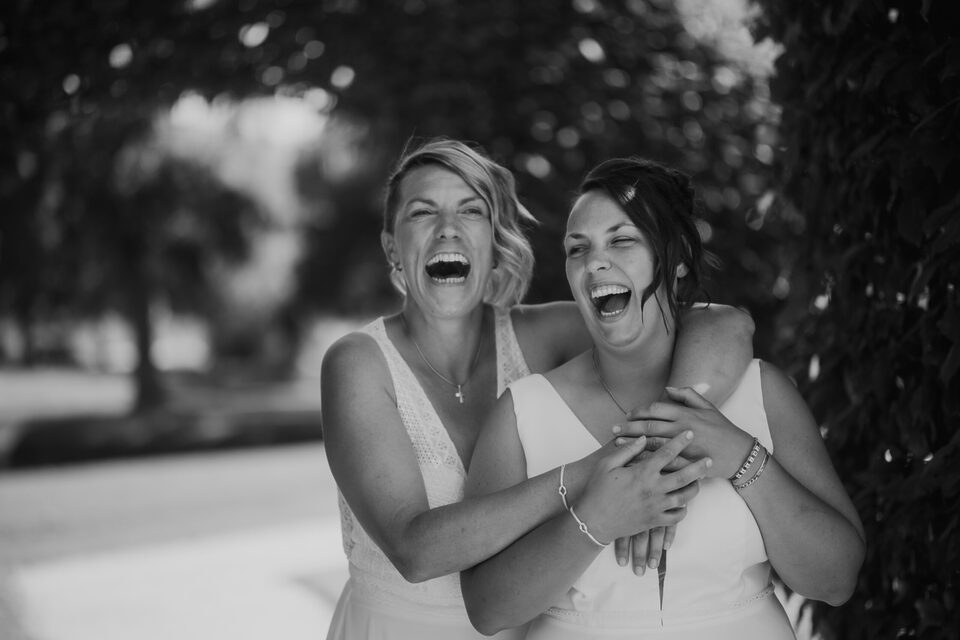 Hochzeitsfotografie von Patricia Bakker: Zwei Bräute lachen herzlich, eine umarmt die andere – authentischer Moment (Schwarzweiß).