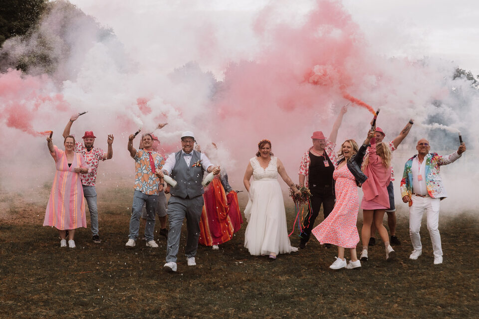 Brautpaar und Gäste laufen jubelnd durch rosafarbenen Rauch auf einer Festivalhochzeit im Freien - perfekt für alle, die alternativ heiraten.