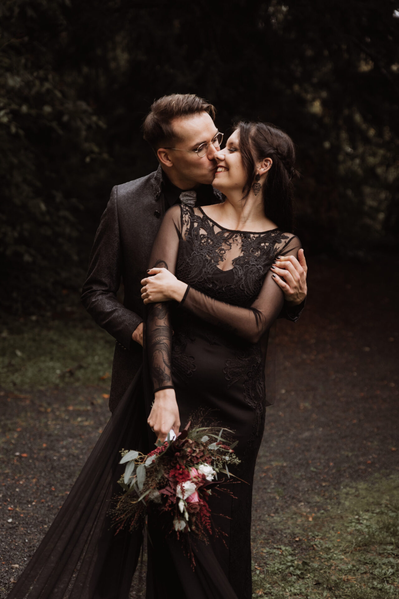Intimer Kuss im Wald, festgehalten von Patricia Bakker – Braut in schwarzer Spitze mit Strauß, Dark-Romance Paarshooting.