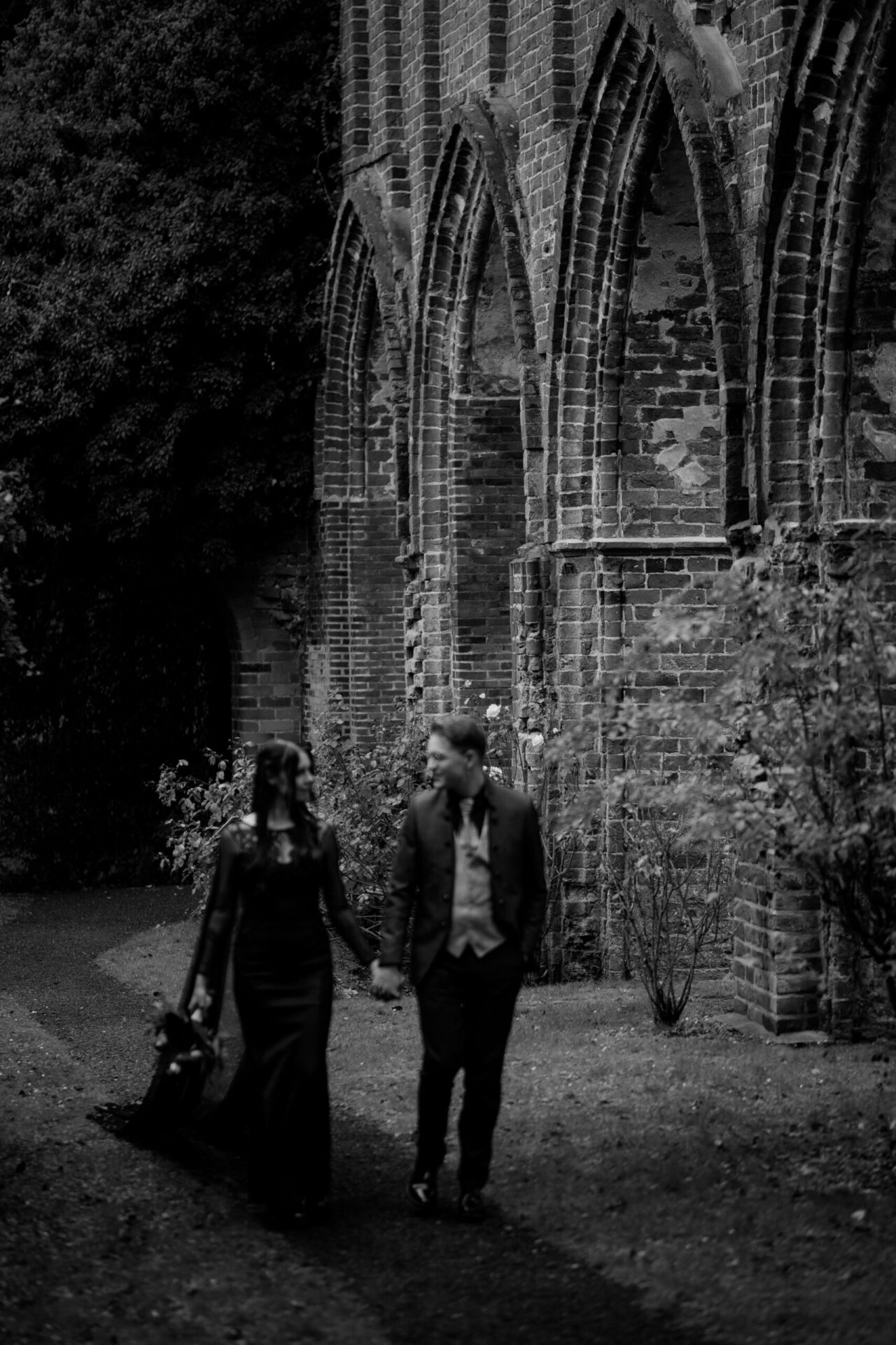 Schwarzweiß Foto eines Hochzeitspaares, spazierend an Backsteinbögen entlang – moody Paarshooting.