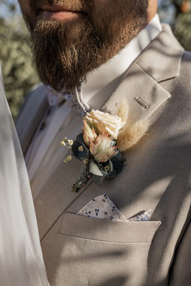 Floraler Hochzeitsanstecker mit cremefarbener Rose, Protea, Eukalyptus und Gräsern an einem grauen Anzug