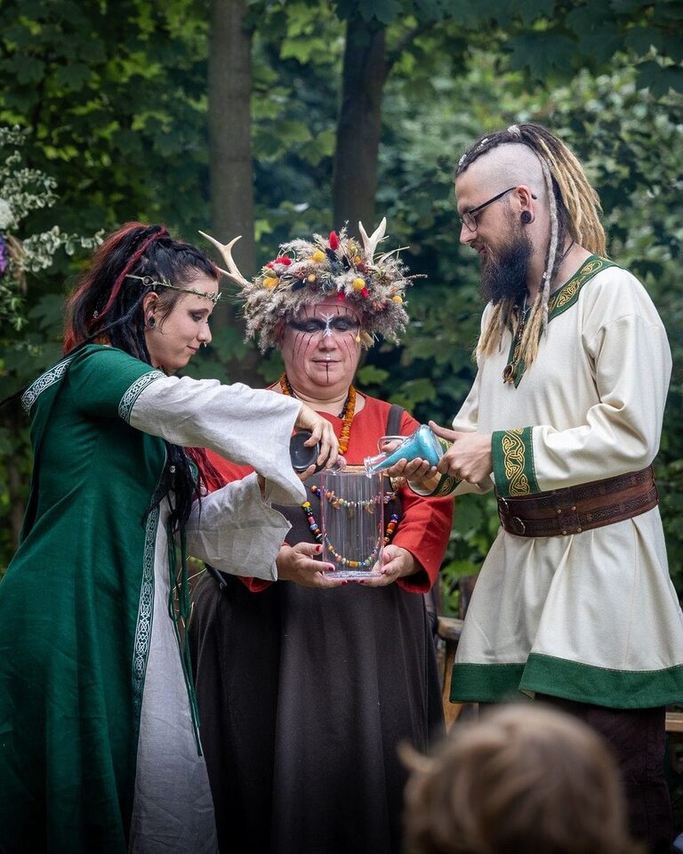 Ein alternativ gekleidetes Hochzeitspaar führt gemeinsam mit der freien Rednerin Nicole ein Ritual bei seiner Wikingerhochzeit durch. Braut und Bräutigam gießen gleichzeitig Flüssigkeit aus zwei Gefäßen in einen großen transparenten Krug, den Nicole zwischen ihnen hält. Die Zeremonie findet draußen zwischen Bäumen statt.