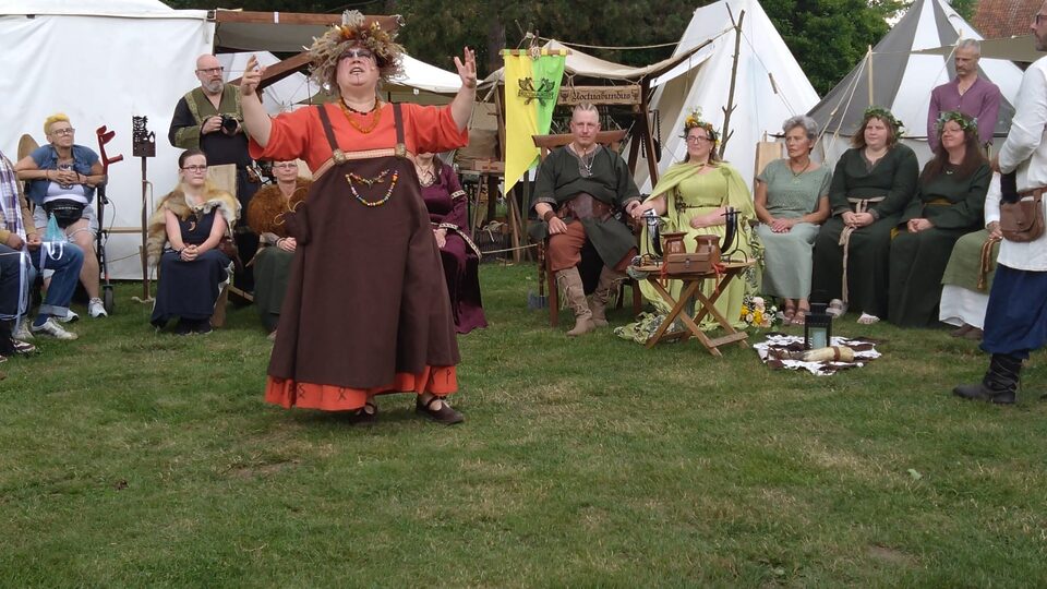 Freie Rednerin Nicole steht mit erhobenen Armen im Mittelpunkt einer Wikingerhochzeit unter freiem Himmel. Die Hochzeitsgesellschaft sitzt im Halbkreis um sie herum; im Hintergrund stehen große weiße historische Zelte und weitere Elemente eines Wikingerlagers. Nicole trägt ihr rotbraunes Gewand und einen Kopfschmuck aus Naturmaterialien.